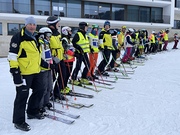 Auf dem Bild steht eine große Gruppe von Skifahrerinnen und Skifahrern nebeneinander auf einer schneebedeckten Fläche. Sie sind in einer langen Reihe aufgestellt und tragen alle Ski an den Füßen sowie Skistöcke in den Händen. Viele Personen tragen le