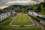 Das Bild zeigt von oben fotografiert den Platz des Geschehens, den Kunstrasenplatz unmittelbar vor dem Hauptgebäude des Sportzentrums. Es zeigt die beiden Mannschaften in Aktion und die Zuseher:innen am Rand des Platzes