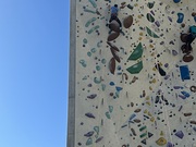 Auf dem Bild ist eine hohe Outdoor-Kletterwand zu sehen, übersät mit bunten Klettergriffen in verschiedenen Formen und Farben. Lilli ist gerade gesichert am Klettern und bewegen sich die Wand hinauf. Links daneben spannt sich ein klarer, wolkenloser 