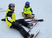 Auf dem Bild sitzen zwei Personen im Schnee, beide mit Skiern an den Füßen. Links sitzt eine erwachsene Begleitperson in gelb-schwarzer Skijacke und Helm, rechts daneben ein Kind mit blauem Helm und gelber Weste mit der Aufschrift „BLIND“. Beide mach
