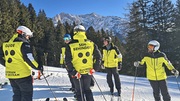 Das Bild zeigt eine Gruppe von Skifahrerinnen auf einem verschneiten Hang, umgeben von Tannen und Bergen. Die Personen tragen auffällige gelb-schwarze Jacken mit Aufschriften wie „GUIDE“ und „SEHBEHINDERT oder BLIND“, sowie „AUSTRIAN BLIND SKITEAM“ u