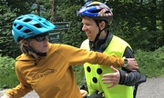 Auf dem Bild stehen zwei Personen mit Fahrradhelmen mit ihrem Tandem auf einem Waldweg. Die Frau links trägt einen blauen Helm und einen gelben Hoodie, während sie ihren Arm schützend oder unterstützend vor den Mann hinter ihr hält. Der Mann trägt ei